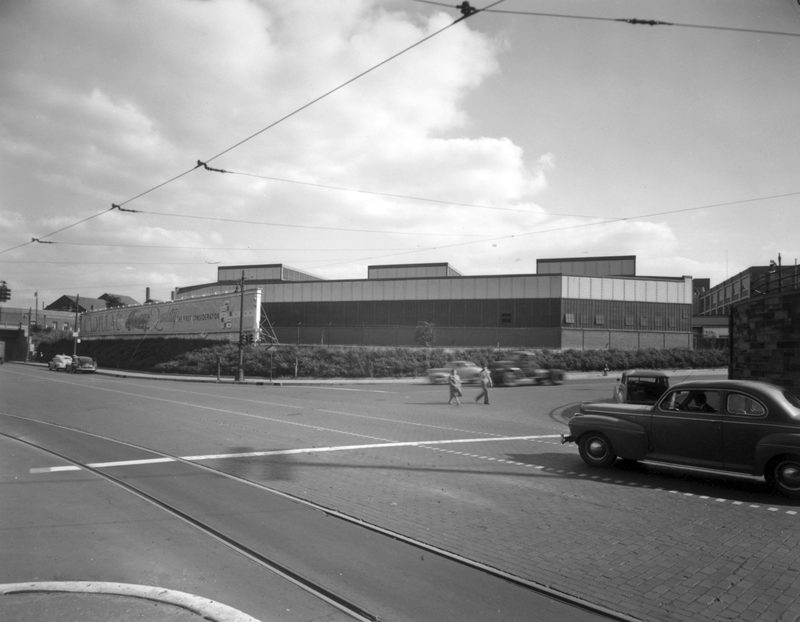 Vintage black-and-white street view of Cadillac's Clark Street campus in Detroit, with a period automobile and streetcar tracks in the foreground